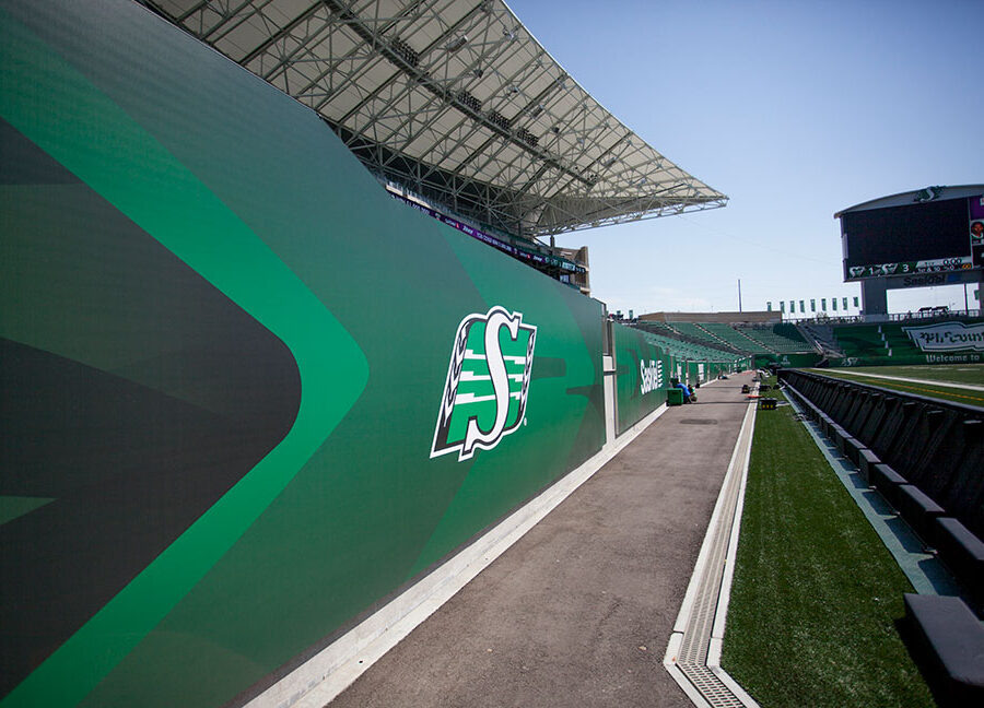 "Sideline view of a stadium with green Saskatchewan Roughriders branding, empty seats, and a scoreboard under a clear sky."