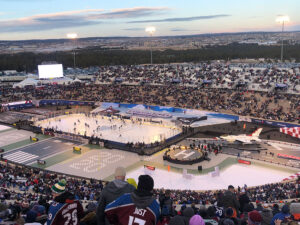 An outdoor NHL Stadium Series hockey game is played in a packed stadium featuring an airfield-themed field with a parked fighter jet.