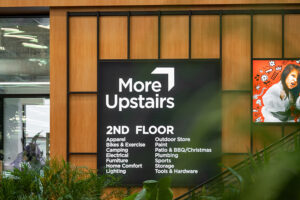 "A black ""More Upstairs"" sign with an upward arrow points to the 2nd floor, listing departments like ""Apparel"" and ""Outdoor Store"", hanging on a wood-paneled wall next to a red poster of a child in a karate gi, with green plants in the foreground."