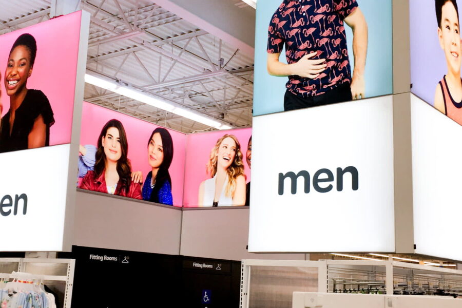 Brightly lit overhead signs with diverse models mark the 'women', 'men', and 'kids' clothing sections of a retail store.