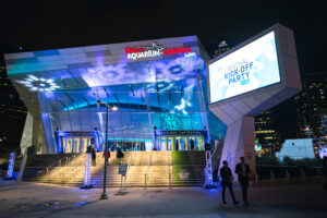 "Ripley's Aquarium of Canada glows with blue and purple lights at night, featuring a large screen promoting a ""Festival Kick-Off Party"" and ""104th Grey Cup Festival"" banners."