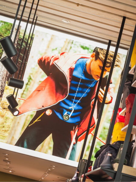 Outdoor apparel store display featuring a large image of a man in a red jacket with a compass, flanked by shelves of colorful bags and Hunter boots.