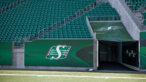 "An empty stadium features green seats, a football field, and Saskatchewan Roughriders branding on a wall and player tunnel."