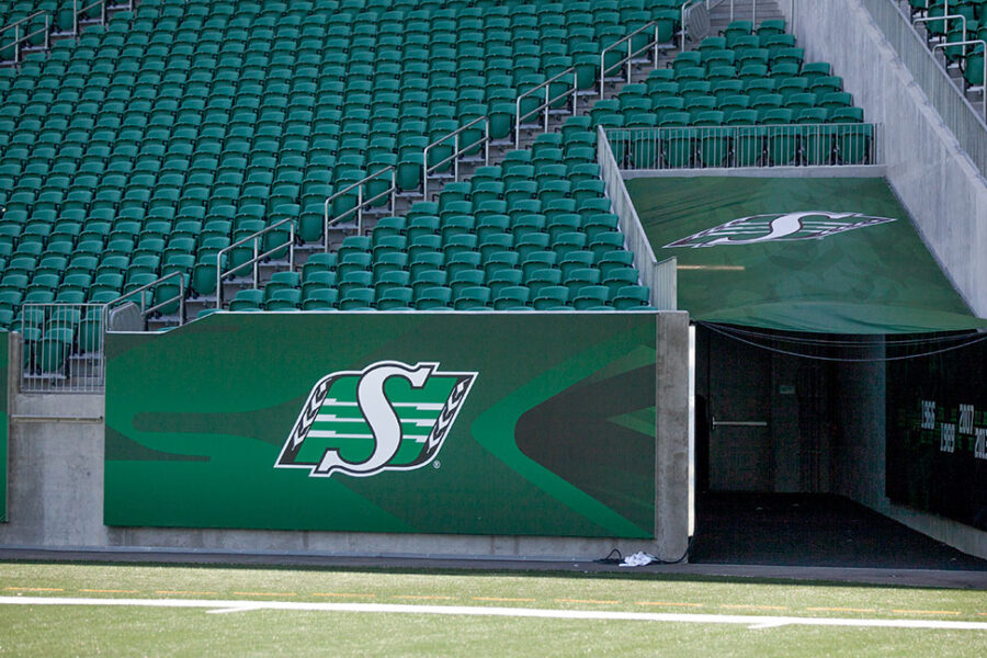"An empty stadium features green seats, a football field, and Saskatchewan Roughriders branding on a wall and player tunnel."