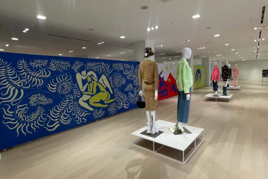 Several fashion mannequins are displayed in a modern, brightly lit store, with a prominent blue wall mural featuring yellow, Matisse-style figures and patterns.
