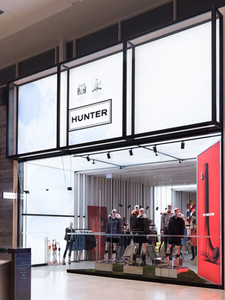 "A Hunter retail store in a modern mall showcases mannequins and a large red boot display, with a Roots store visible adjacent to it."