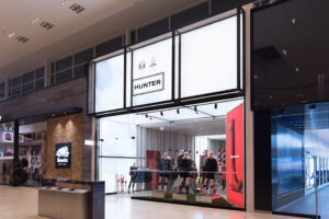 "Hunter store entrance with mannequins and a red boot display, next to a Roots store in a mall."