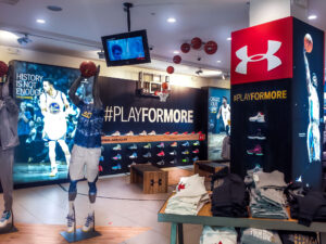 "Under Armour's basketball-themed store display features ""PLAYFORMORE"" signage, Stephen Curry posters, various athletic shoes, and a mini basketball hoop."