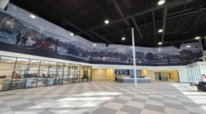 "A modern, spacious interior features a panoramic mural depicting interconnected technological landscapes and silhouettes, with glass-walled offices and a reception area below."