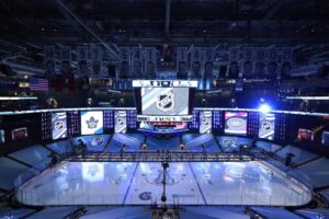 "An empty Scotiabank Arena in Toronto features a brightly lit hockey rink with the Stanley Cup 2020 logo, a large NHL scoreboard, and retired player banners hanging overhead."