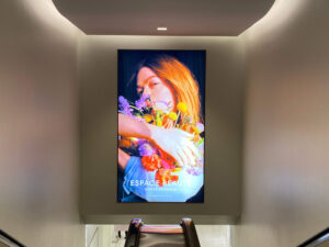 "A large digital advertisement for ""Espace Beaute,"" featuring a woman holding colorful flowers, is prominently displayed above escalators."