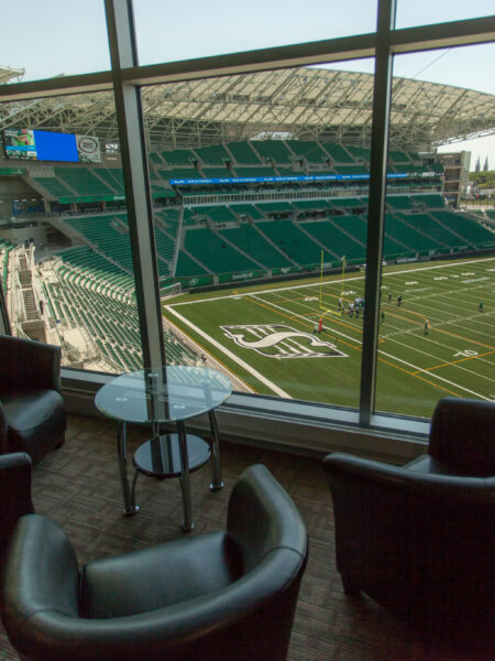A comfortable lounge area with dark armchairs and a glass table overlooks a football stadium with a green field, a few players, and rows of empty green seats.