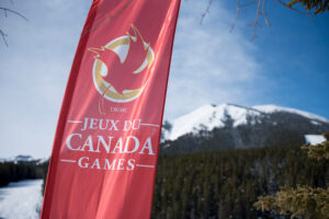 "A red ""Jeux du Canada Games"" flag stands prominently against a backdrop of snowy mountains under a clear blue sky."