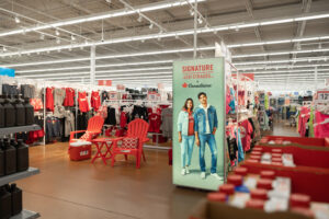 "A retail store aisle features a large Levi Strauss x Canadiana promotional sign, red clothing displays, and matching red Adirondack chairs."