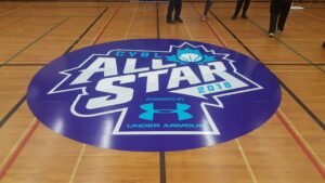 A purple and white CYBL All Star 2018 logo by Under Armour is displayed on a wooden gymnasium floor.