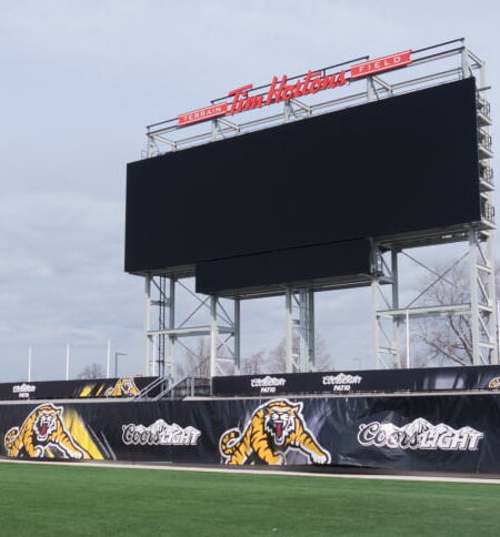 "A wide shot of Tim Hortons Field stadium featuring a large black scoreboard, green playing field, and black-and-yellow tiger-themed banners."