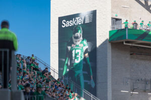 A large SaskTel banner featuring a football player in a green #13 jersey overlooks stadium seating filled with green-clad fans.