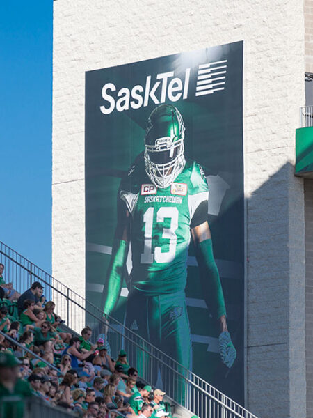 A large SaskTel banner featuring a football player in a green #13 jersey overlooks stadium seating filled with green-clad fans.