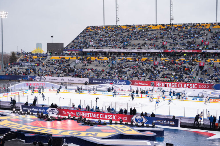"The Toronto Maple Leafs and Buffalo Sabres play an outdoor hockey game at the Tim Hortons Heritage Classic 2022 in Hamilton, with a large crowd filling the stadium stands."
