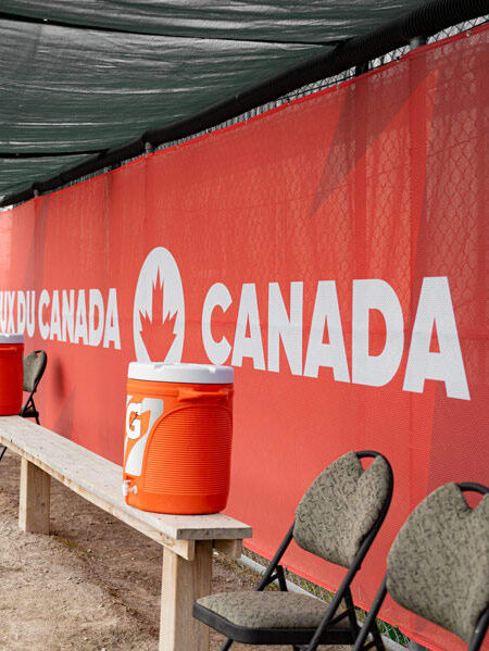 A sports dugout features a wooden bench, orange coolers, and chairs against a red banner for the Canada Games.