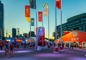 "An outdoor World Cup Hockey 2016 Fan Village at dusk, featuring national flags, event tents, and crowds against a city skyline."
