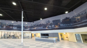 "A modern lobby features a panoramic mural of industrial landscapes overlaid with abstract tech graphics, a central reception desk, and a checkered floor."