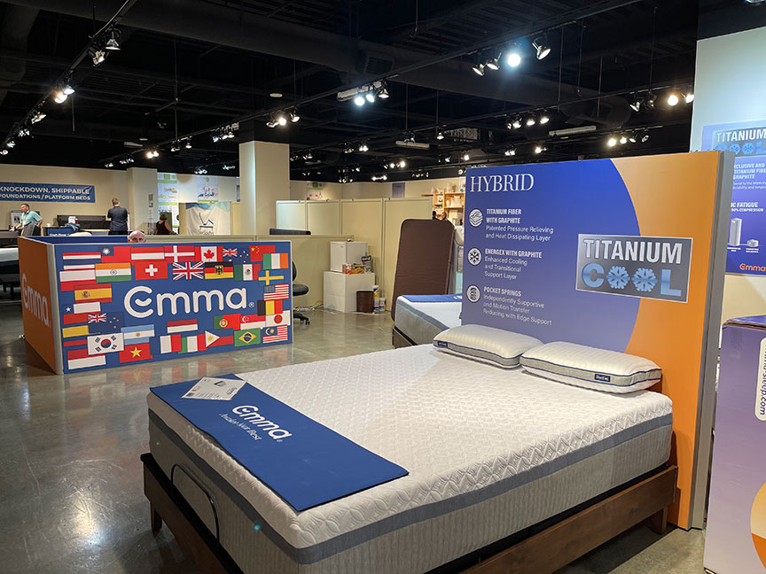 A mattress showroom featuring Emma brand displays, including a ""Titanium Cool Hybrid"" mattress and a large sign with international flags.