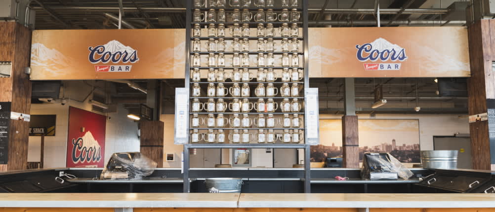An empty Coors bar features two large Coors signs, a wooden counter, and a tall central display of glass mugs.