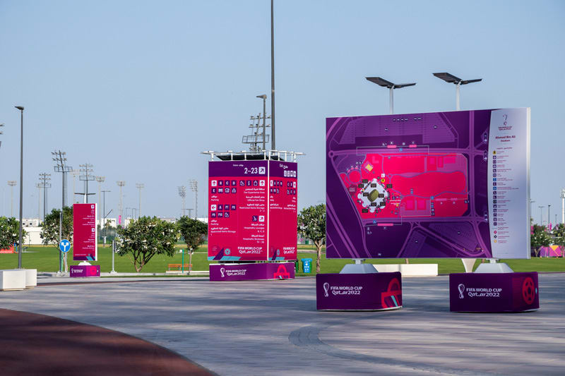 Outdoor plaza featuring large purple and pink FIFA World Cup Qatar 2022 directional signs and a stadium area map.