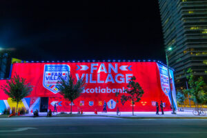 "A large, brightly lit red banner for the ""World Cup of Hockey 2016 Fan Village Scotiabank"" stands on a city street at night, with a few people and trees visible."