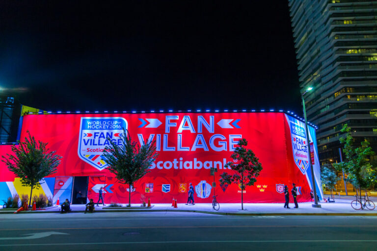 "A large, brightly lit red banner for the ""World Cup of Hockey 2016 Fan Village Scotiabank"" stands on a city street at night, with a few people and trees visible."