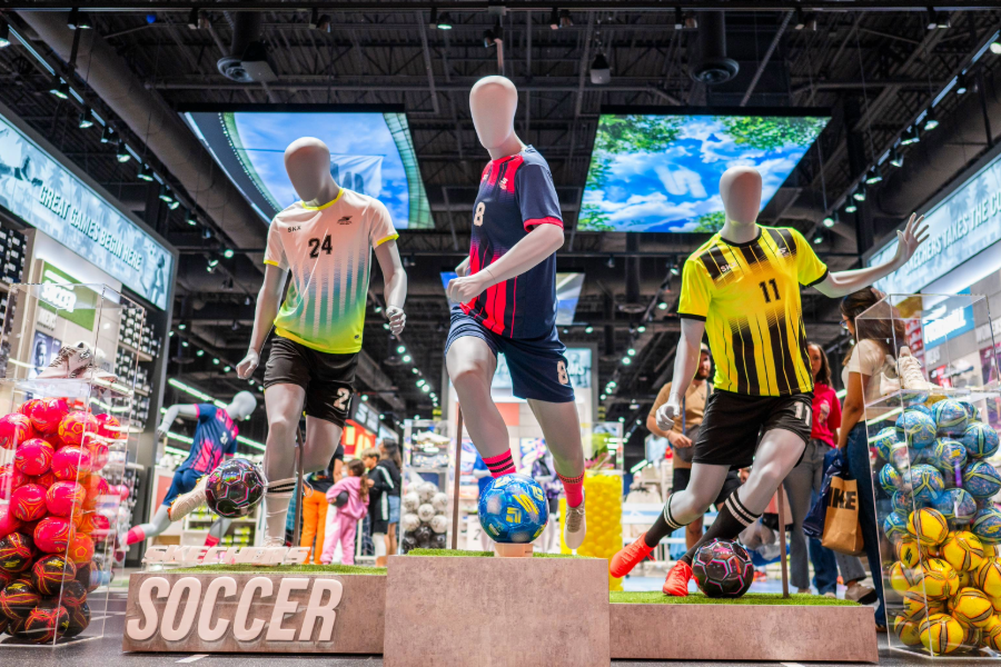 A vibrant soccer display in a sports store features mannequins in action poses surrounded by soccer balls, uniforms, and footwear.