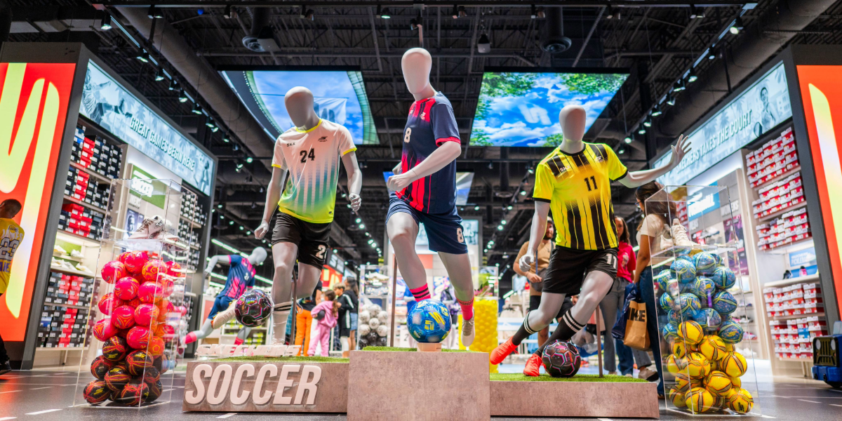 A vibrant soccer display in a sports store features mannequins in action poses surrounded by soccer balls, uniforms, and footwear.