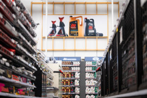 "A wide shot down a store aisle featuring shelves stocked with automotive products, topped by a large banner displaying jack stands and motor oil bottles."