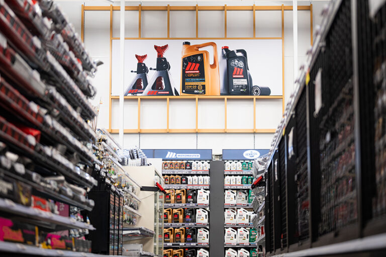 "A wide shot down a store aisle featuring shelves stocked with automotive products, topped by a large banner displaying jack stands and motor oil bottles."