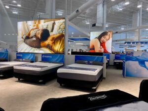 "A brightly lit mattress showroom features multiple beds, large digital screens displaying sleeping people, and ""Rene by Rooms To Go"" branding."