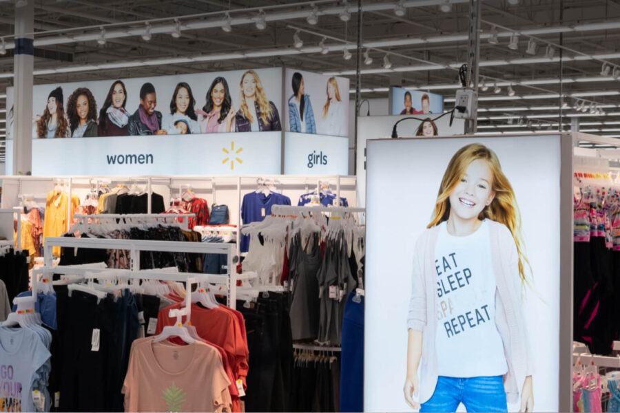 A Walmart clothing department features racks of women's and girls' apparel, with overhead signs displaying models and a large illuminated poster of a smiling girl.