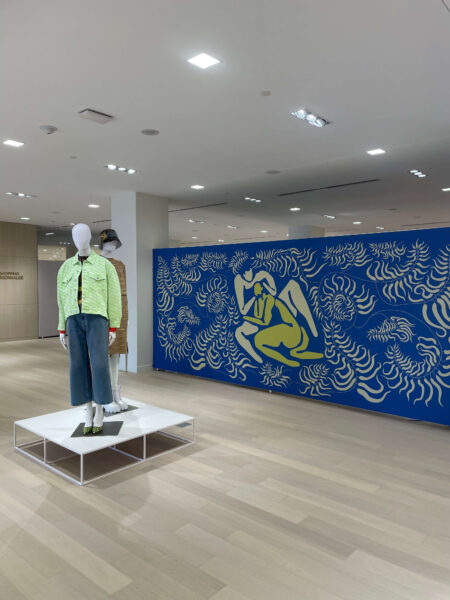 A bright retail space features two mannequins displaying modern clothing and a large blue wall with a yellow and cream abstract mural.