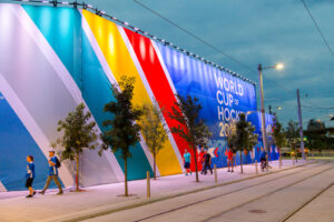 "A large banner advertising the ""WORLD CUP OF HOCKEY 2016"" with colorful diagonal stripes covers a building facade as people walk along a city street with tram tracks."