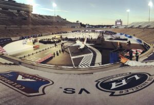 "An outdoor hockey rink in a stadium features a white fighter jet on an aviation-themed runway surrounding the ice, with Avalanche and Kings logos in the foreground."