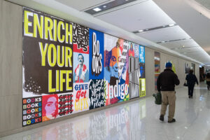 "A vibrant, pop-art style collage mural with phrases like ""ENRICH YOUR LIFE"" and ""BOOKS"" spans a bright modern hallway where people walk past."