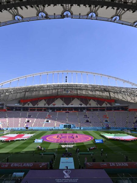 A wide shot of a modern stadium with a green soccer field, featuring the English and Iranian flags laid out and FIFA World Cup Qatar 2022 branding.