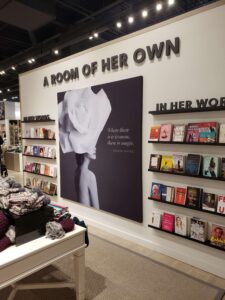 "A women's literature display features a large ""A ROOM OF HER OWN"" sign, a black-and-white poster of a woman with a white rose and the quote ""Where there is a woman, there is magic,"" and multiple shelves of books labeled ""IN HER WORDS."""