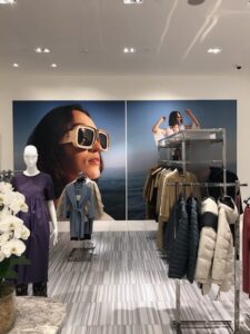 "An apparel store interior featuring two large wall ads of women wearing sunglasses, along with mannequins displaying clothes and racks of outerwear."