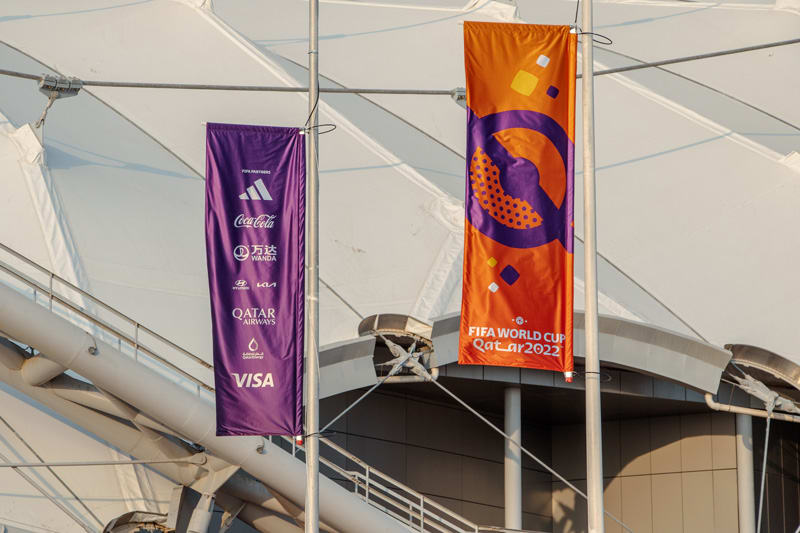 Two banners, one purple listing various sponsors and one orange with the FIFA World Cup Qatar 2022 logo, hang outside a stadium.