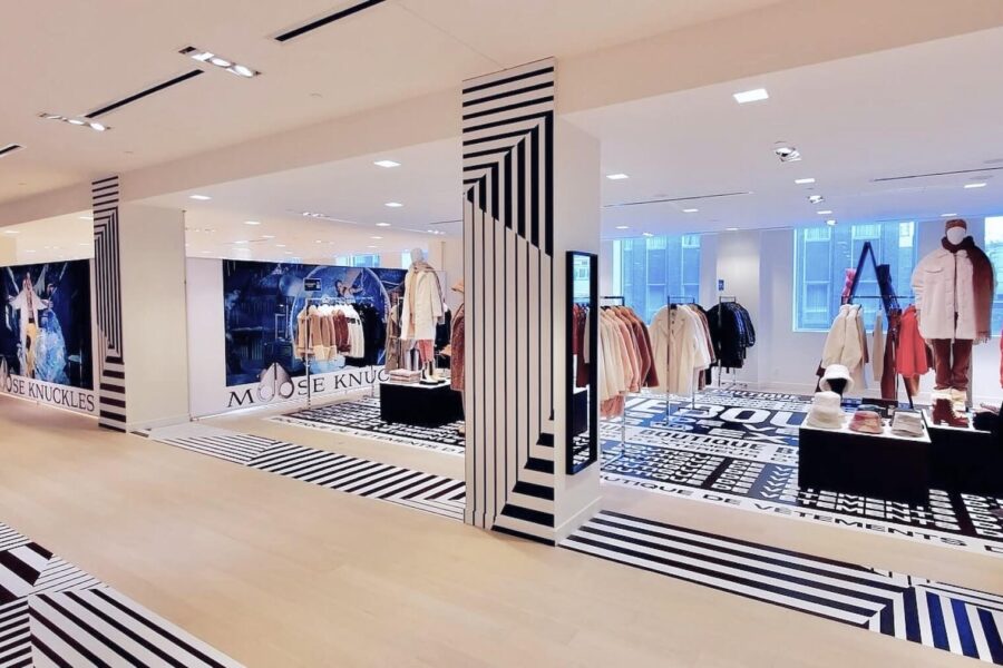 Interior of a Moose Knuckles clothing store with mannequins displaying winter coats and jackets, featuring black and white geometric patterns on the floor and support columns.