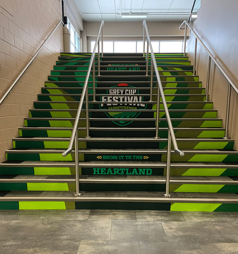 "A staircase decorated in shades of green features ""Grey Cup Festival Saskatchewan"" and ""Bring It To The Heartland"" text on its risers."