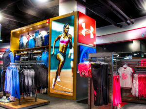"An Under Armour store interior with athletic wear on racks and mannequins, featuring large brand logos and a bright display of a female runner."