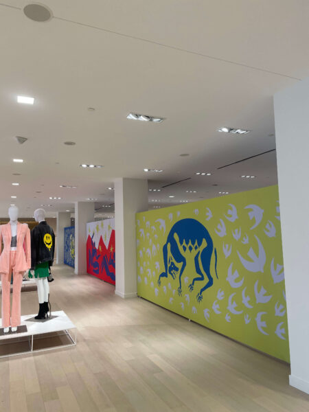 Mannequins in a pink suit and a black smiley-face jacket stand in a store next to walls covered in vibrant graphic art, including a lime green mural with a blue monster and white birds.