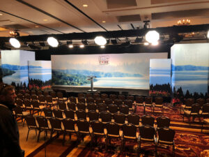 Empty conference room with rows of chairs facing a stage displaying a landscape backdrop and the G7 2018 Charlevoix logo.